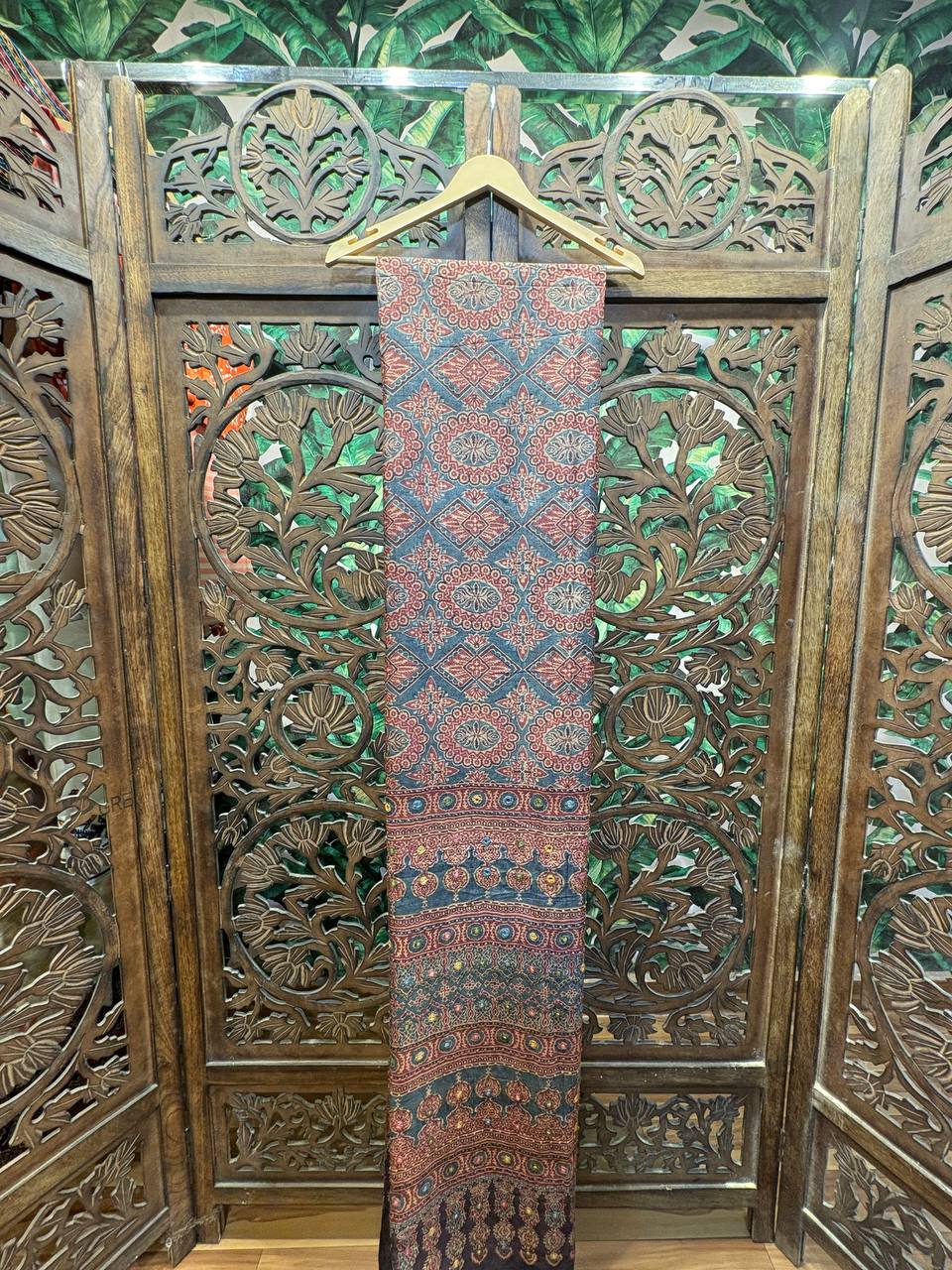 Full length view of the Mihira saree, a teal-blue and maroon Ajrakh saree with Lambani embroidery, hanging against a carved wooden screen.