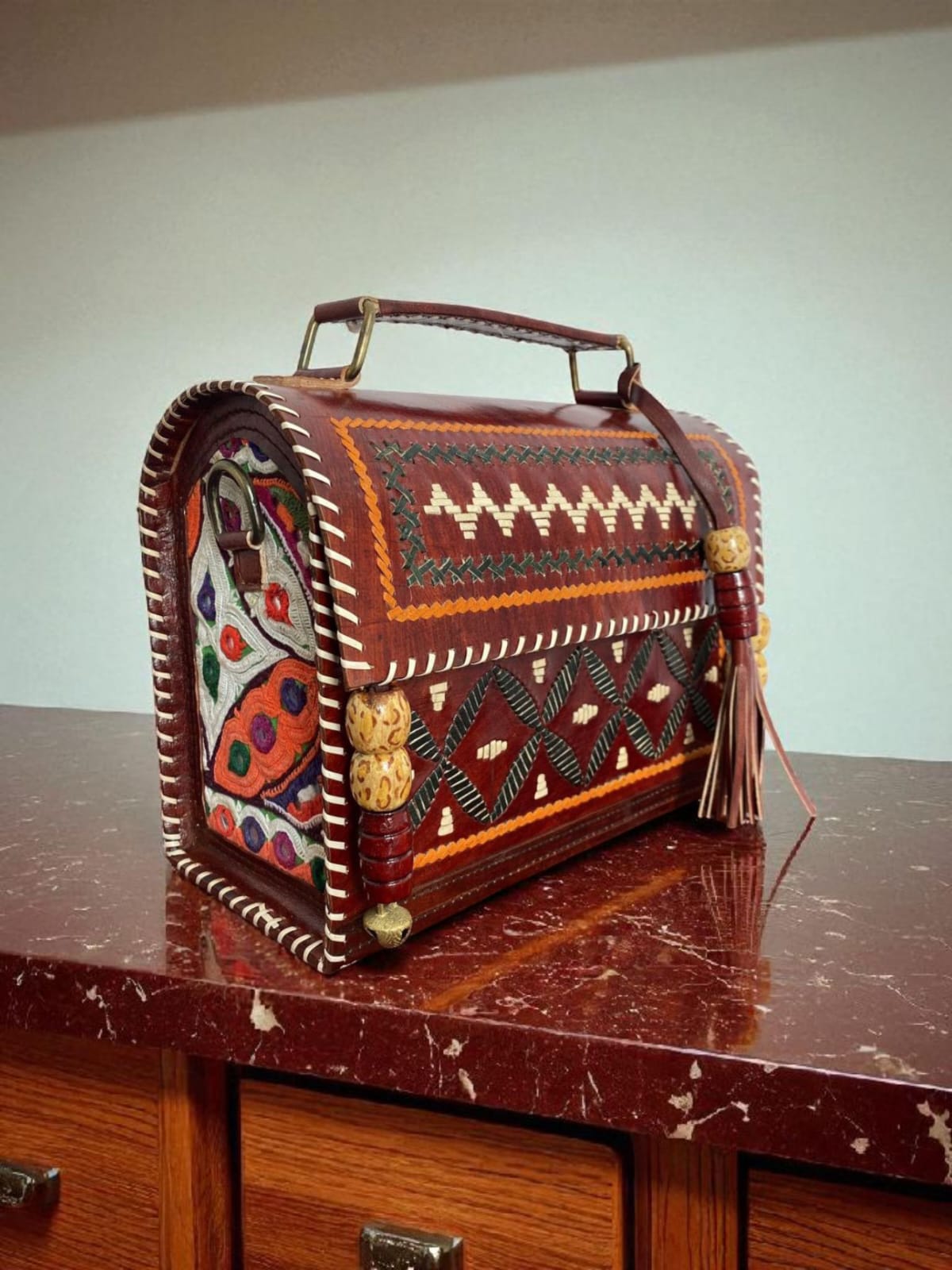 Ethnic handcrafted leather trunk bag with tribal embroidery and a top handle, placed on a carved wooden table.