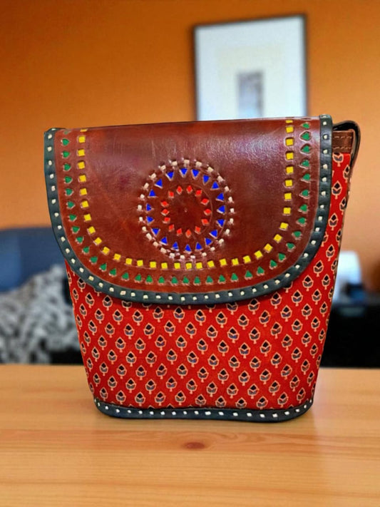 Handcrafted sling bag with a leather flap featuring a colorful Rangoli-inspired design and a red block-printed body.