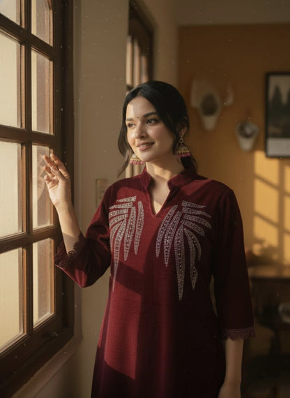 Woman in a maroon embroidered dress standing by a window with warm lighting.